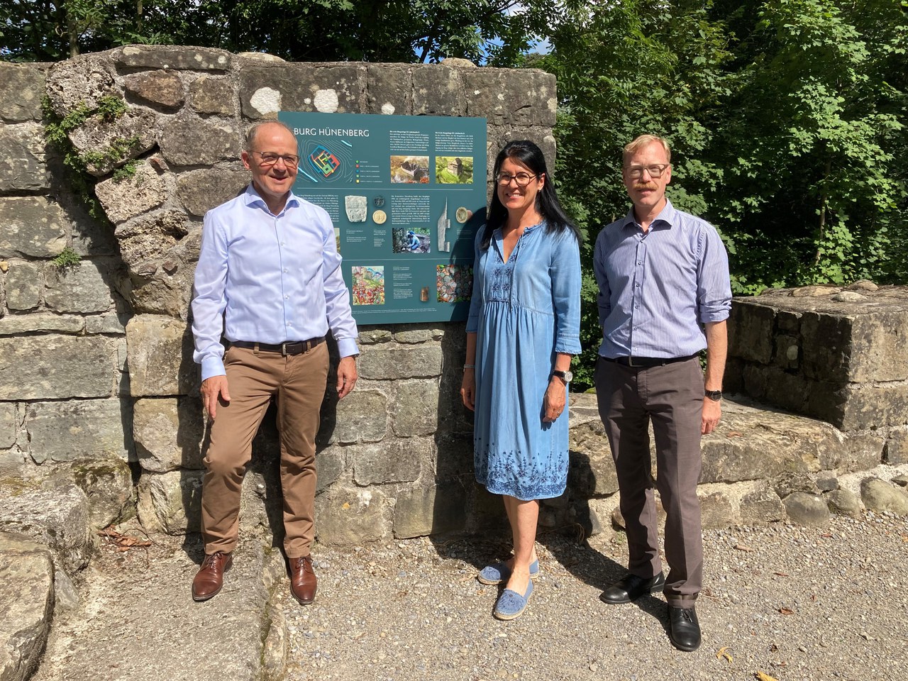 Korporationspräsident Paul Suter, Gemeindepräsidentin Renate Huwyler und Regierungsrat Andreas Hostettler stehen vor der neuen Infotafel bei der Burgruine Hünenberg.