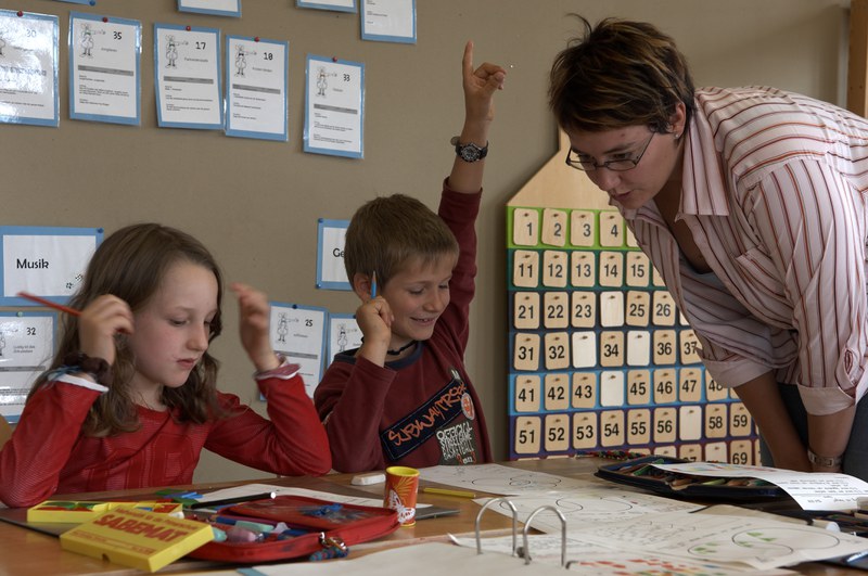 arbeitende Schüler in einem Schulzimmer, welche bei der Lehrperson Hilfe holen